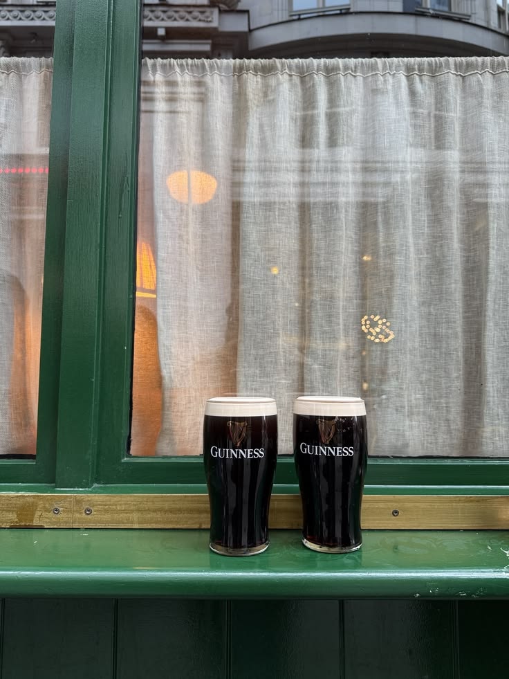Two pints of Guinness on a pub windowsill with green trim and lace curtains.