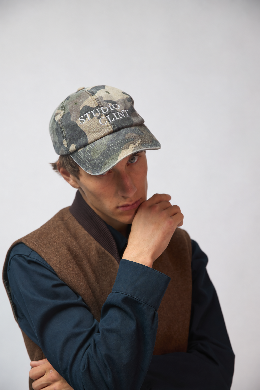 A man wearing a washed camouflage six-panel baseball cap with "Studio Clint" embroidered in white thread across the front, resting his chin on his hand against a light grey studio background.