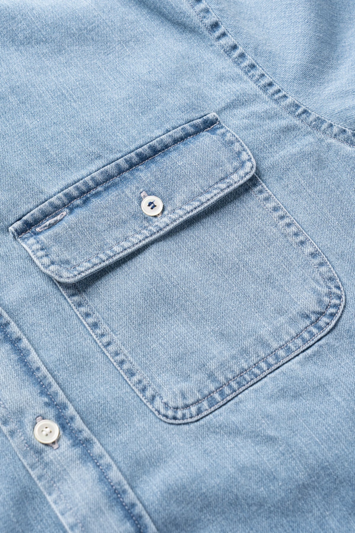 Close-up of a light blue denim shirt showing a buttoned chest pocket and part of the button placket.