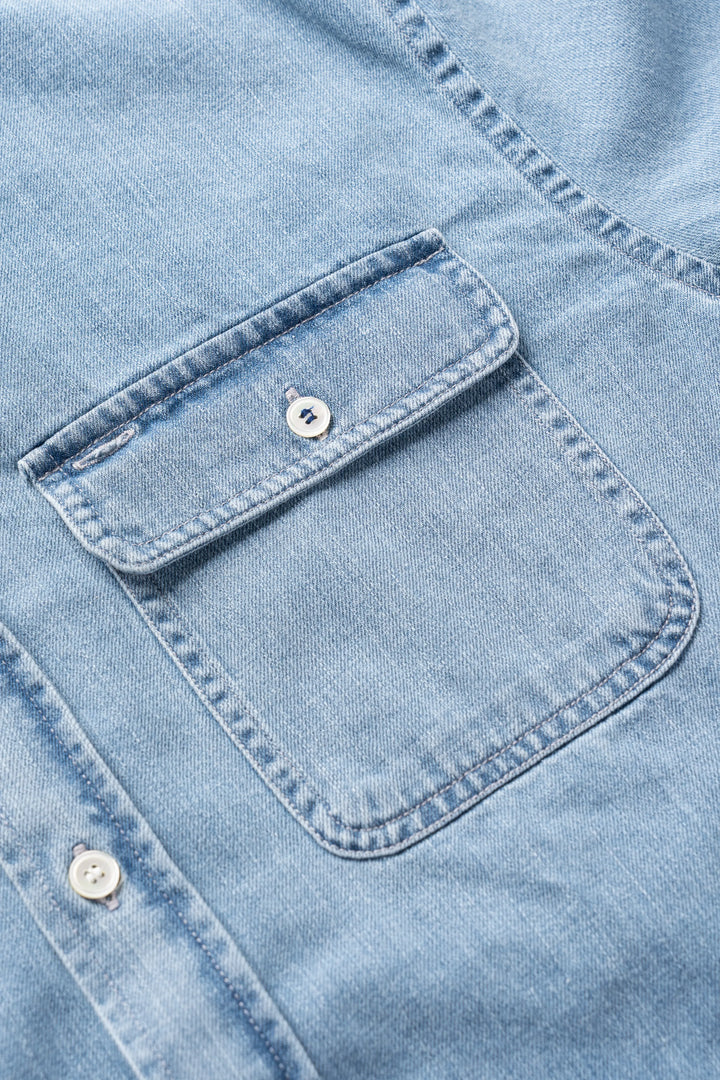 Close-up of a light blue denim shirt showing a buttoned chest pocket and part of the button placket.