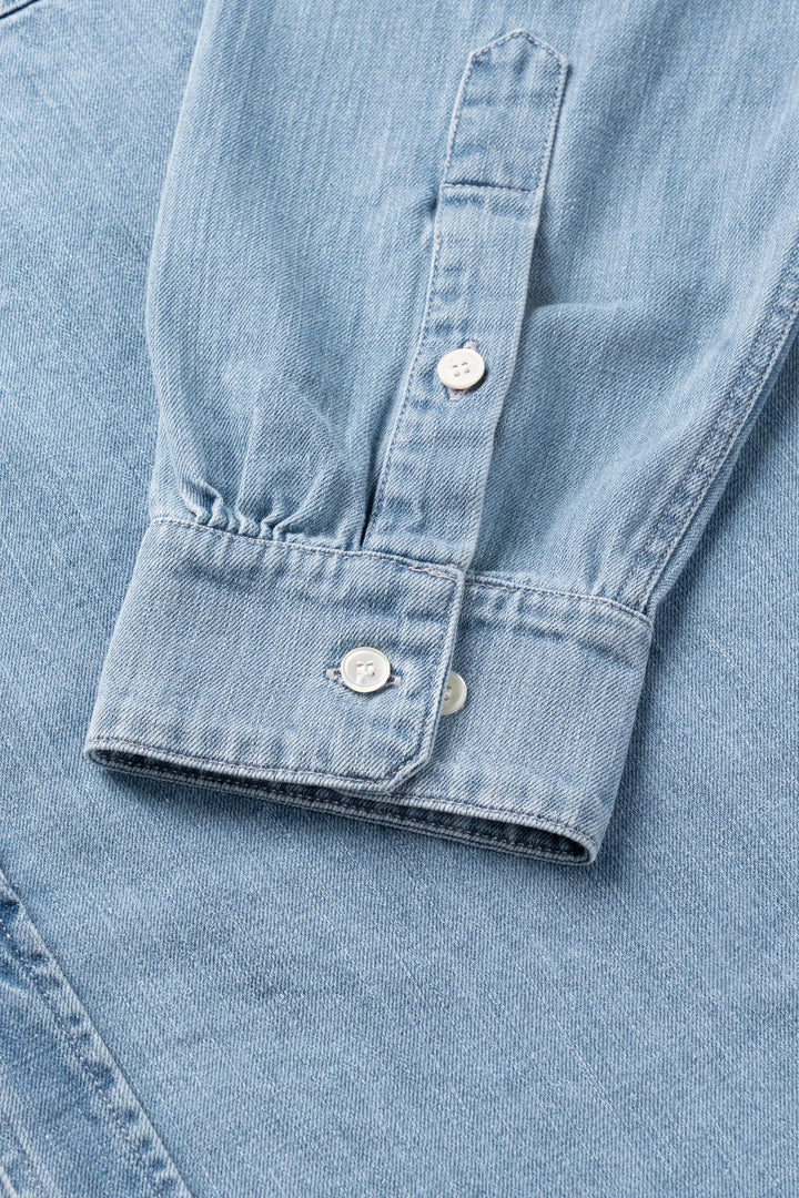 Close-up of a light blue denim shirt sleeve showing the buttoned cuff and front placket.