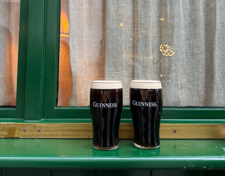 Two Guinness pints on a green ledge with a window and curtain in the background.