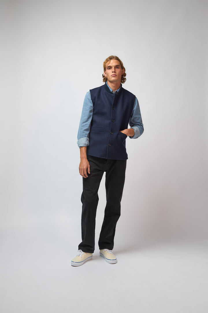 A man wearing a midnight navy loden-wool vest over a navy top, standing against a light grey studio background.
