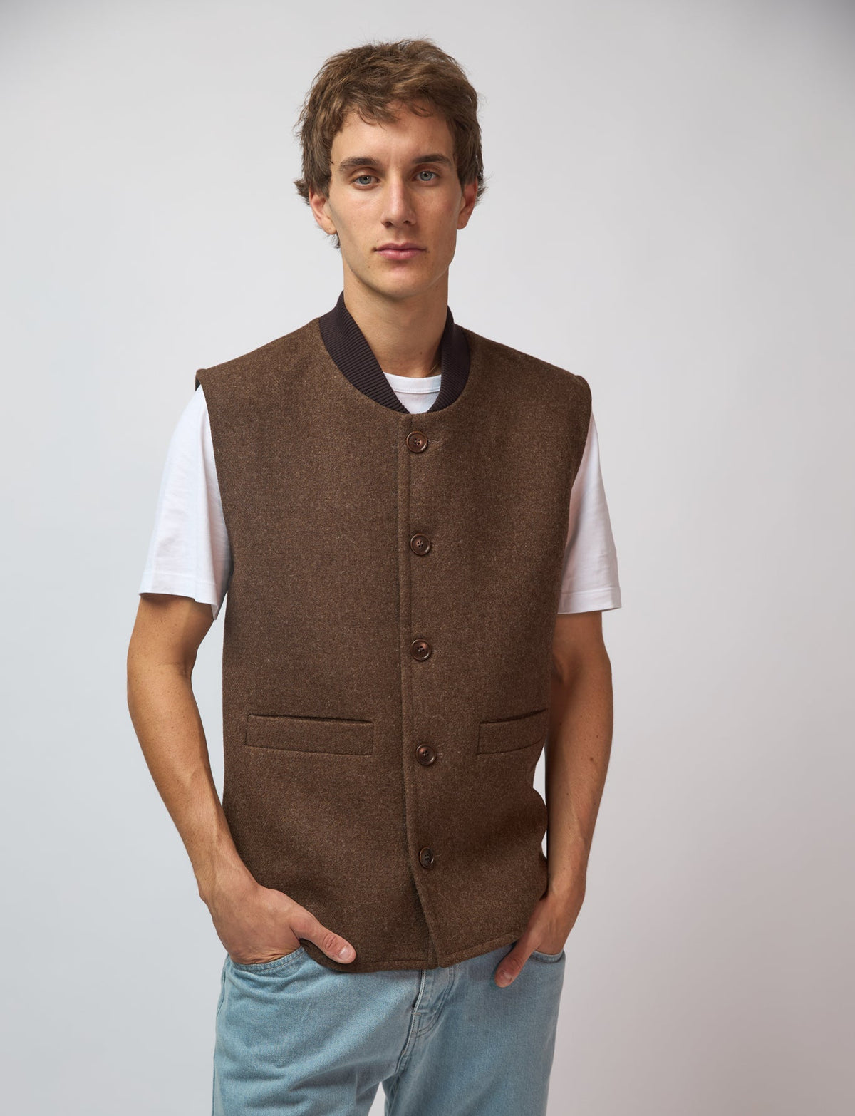 A man wearing a burnt umber loden-wool vest over a navy top, standing against a light grey studio background.