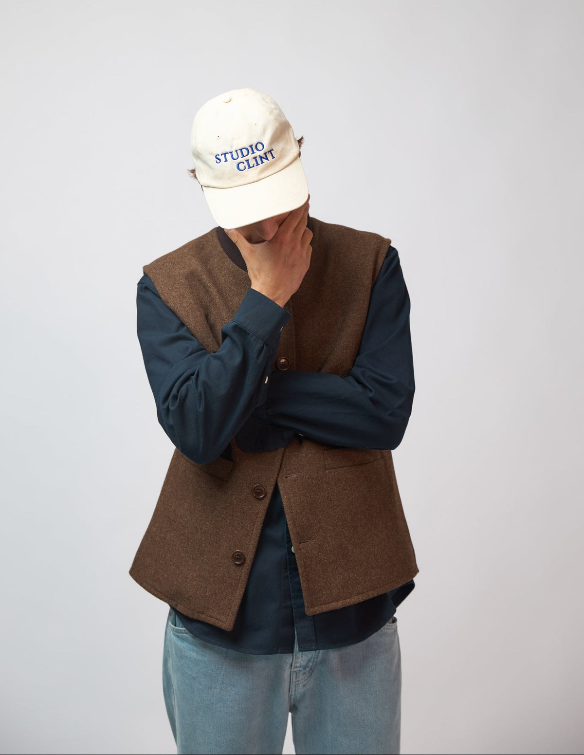 A man wearing a cream baseball cap with "Studio Clint" embroidered in blue thread across the front, touching his hand to his face against a light grey studio background, wearing a brown wool vest over a navy top.