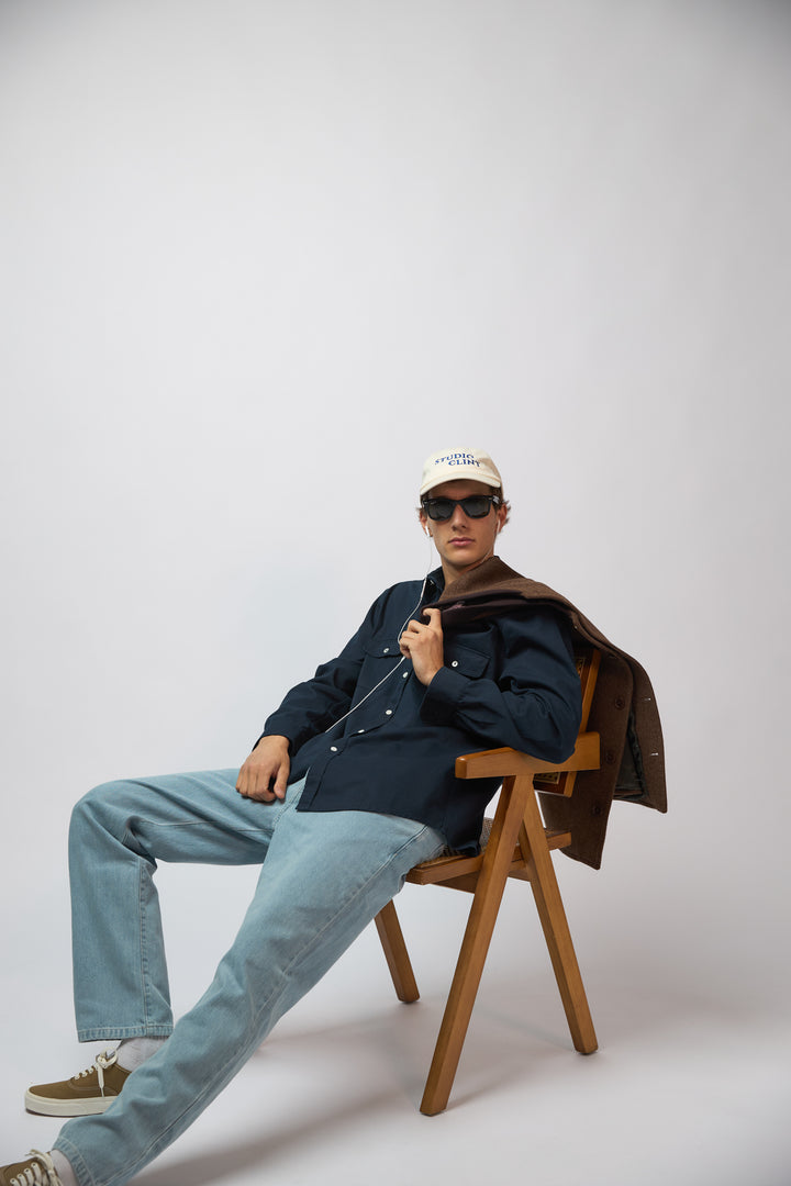 A man wearing a cream baseball cap with "Studio Clint" embroidered in blue thread across the front, shown from a three-quarter angle against a light grey studio background.