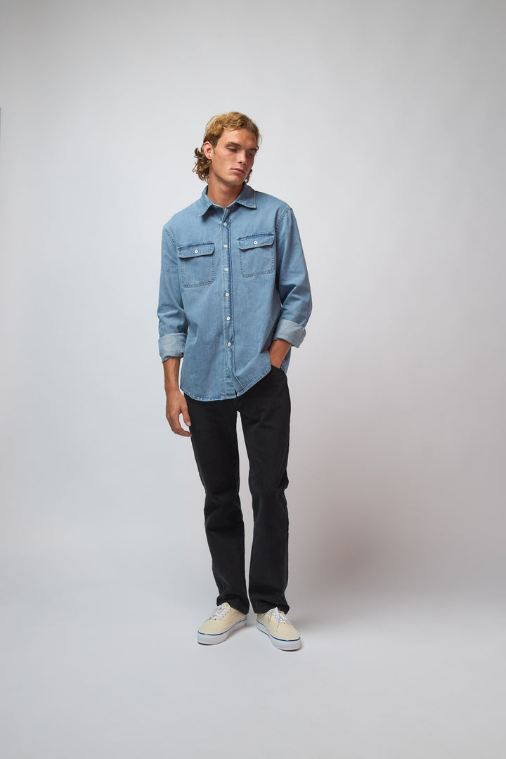 A man wearing the washed denim double pocket shirt layered with denim trousers, standing against a light grey studio background.