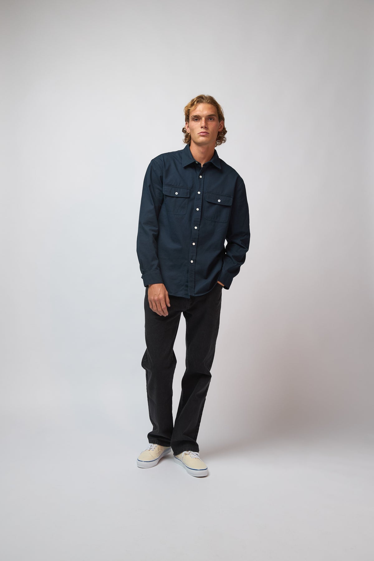 A man wearing the deep navy double pocket shirt with black trousers and cream sneakers, standing against a light grey studio background.