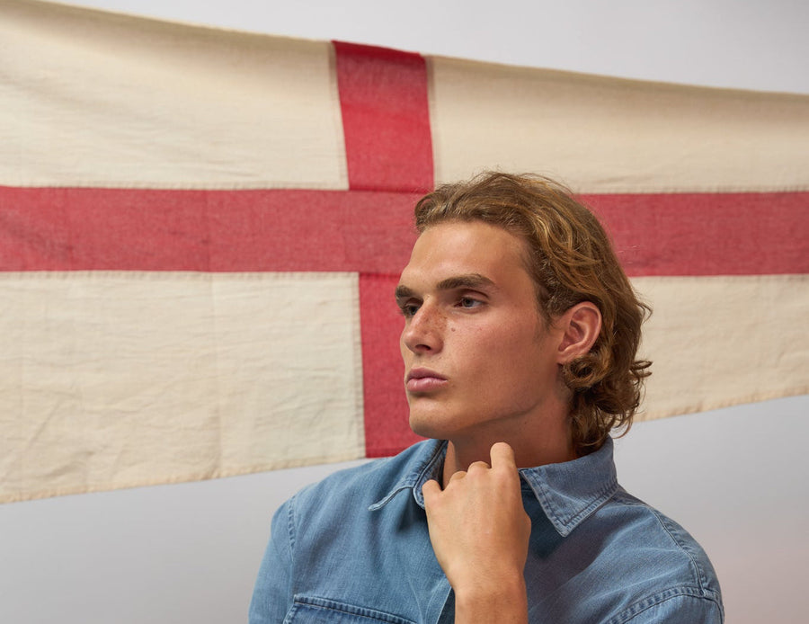 A man wearing a light blue denim button-up shirt and dark pants sits in a wooden chair, looking off-camera against a white wall with a red and cream flag hanging behind him.