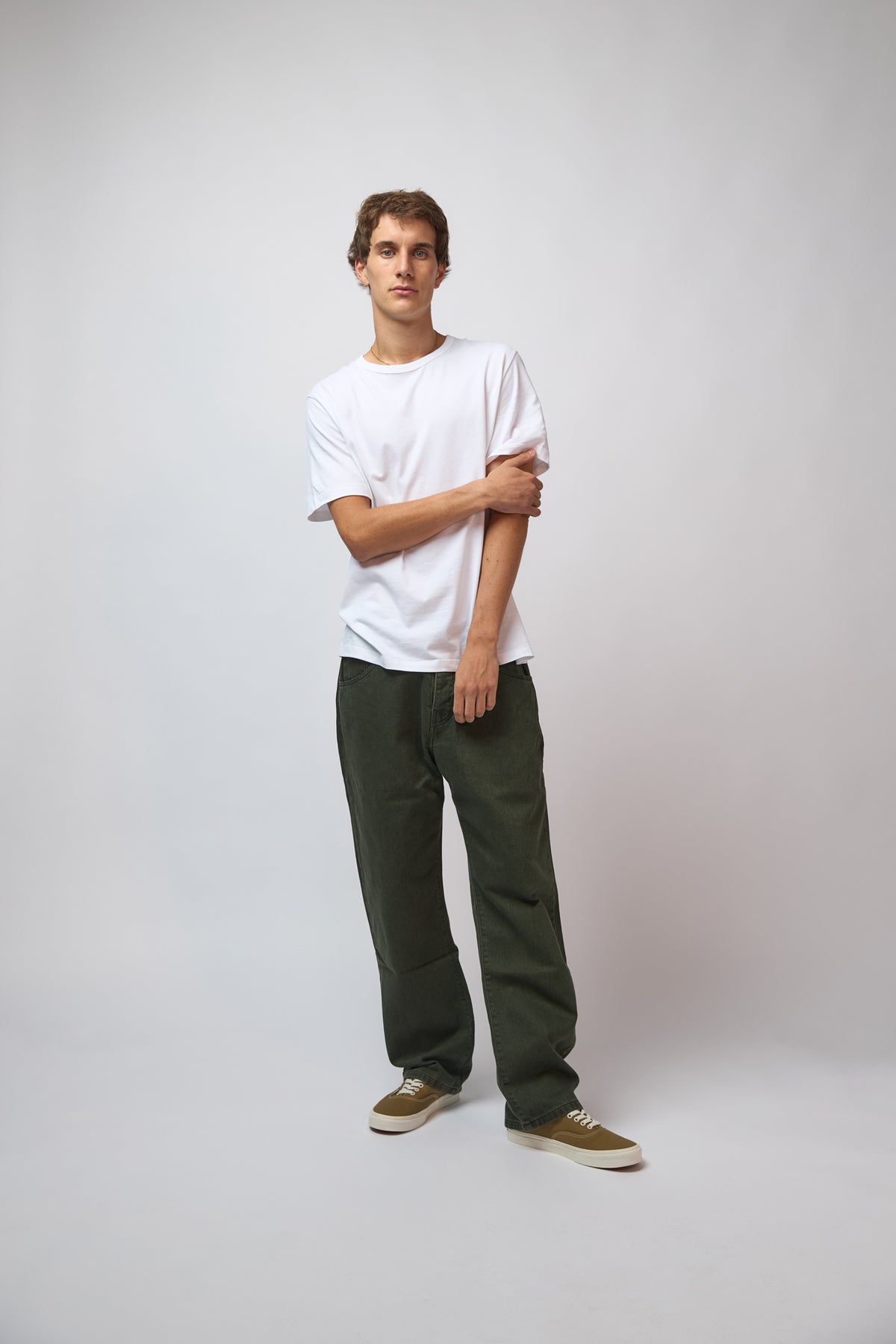 A man modeling a white crewneck t-shirt, dark olive green wide-leg pants, and olive green canvas sneakers, standing against a light gray studio background.