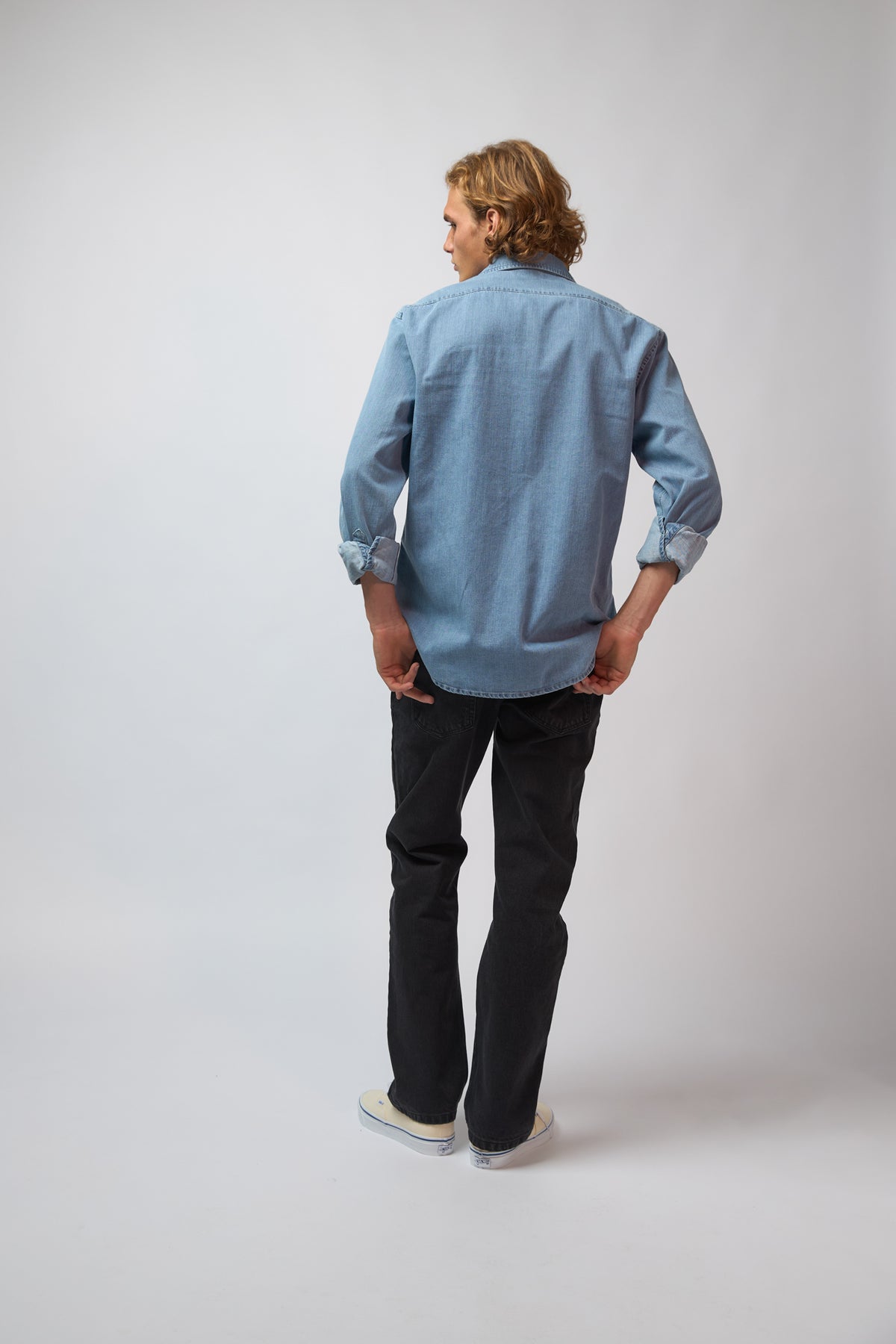 A man wearing the washed denim double pocket shirt, shown from a three-quarter angle against a plain grey studio background.
