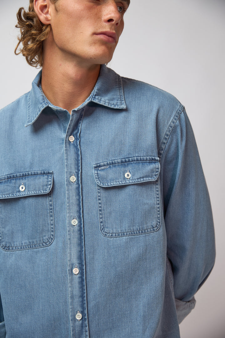 A man wearing the washed denim double pocket shirt with black trousers and cream sneakers, standing against a light grey studio background.