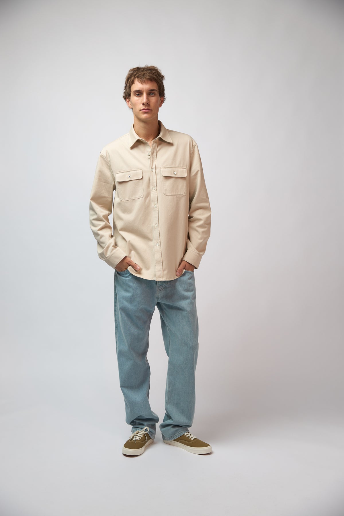 A man wearing the safari-beige double pocket shirt with black trousers and cream sneakers, standing against a light grey studio background.