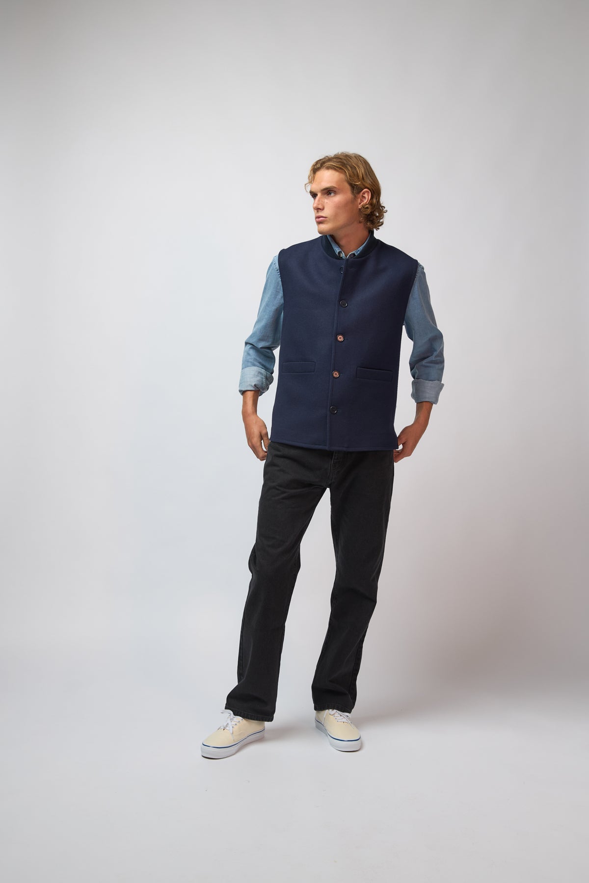 A man wearing a midnight navy loden-wool vest styled with denim trousers, standing against a light grey studio background.
