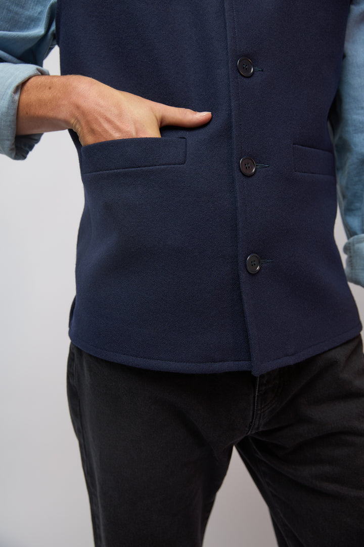 A man wearing a midnight navy loden-wool vest layered over a navy shirt, standing against a plain grey studio background.