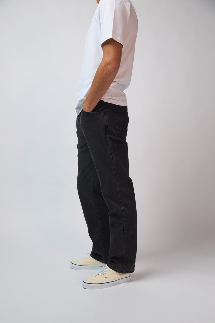 A man wearing coal black denim jeans with a white t-shirt, standing against a light grey studio background.