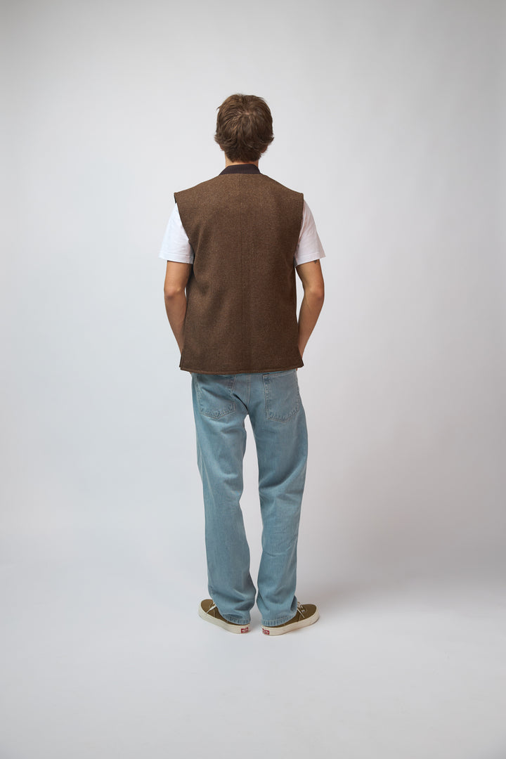 A man wearing a burnt umber loden-wool vest styled with denim trousers, standing against a light grey studio background.
