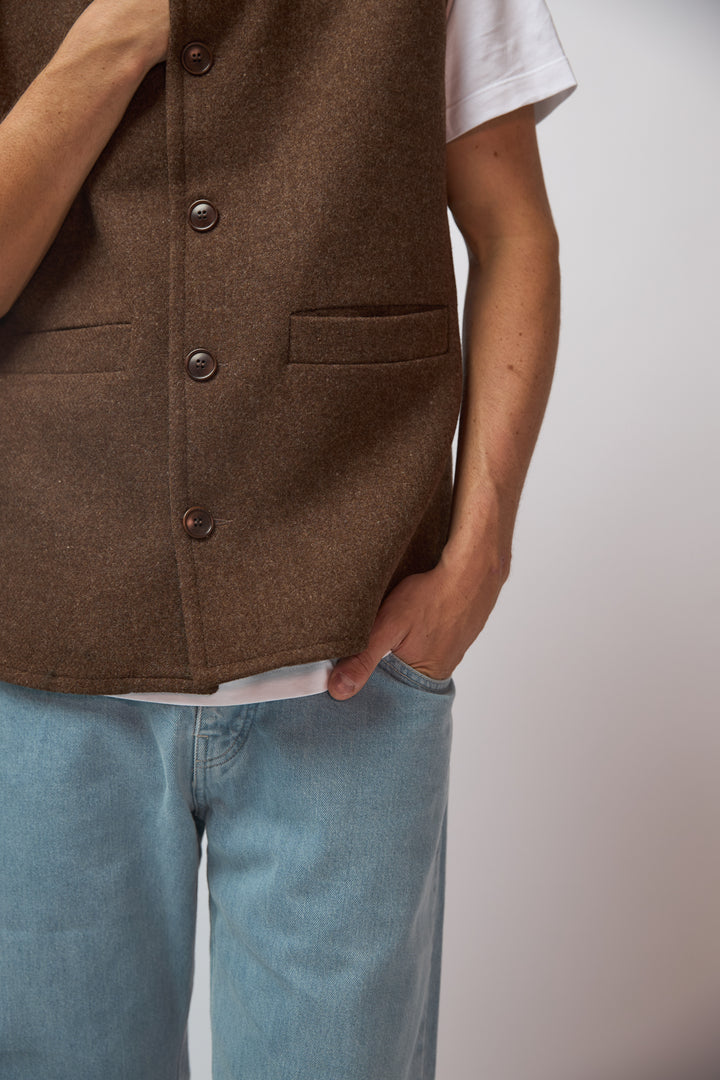 A man wearing a burnt umber loden-wool vest, shown from a three-quarter angle against a plain grey studio background.