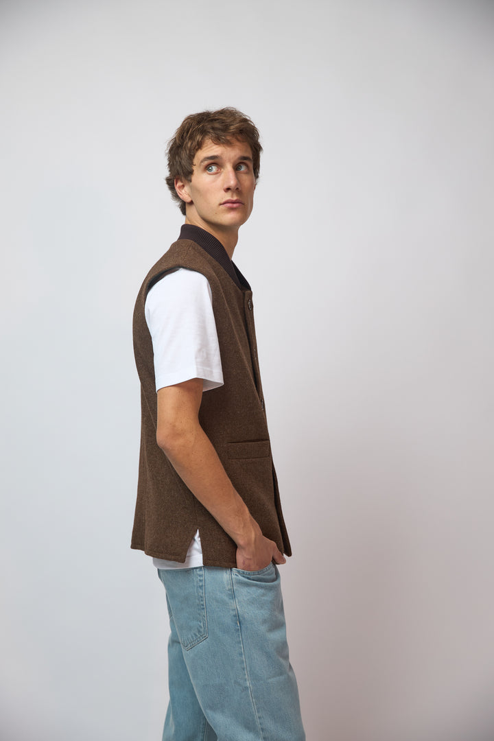 A man wearing a burnt umber loden-wool vest layered over a navy shirt, standing against a plain grey studio background.